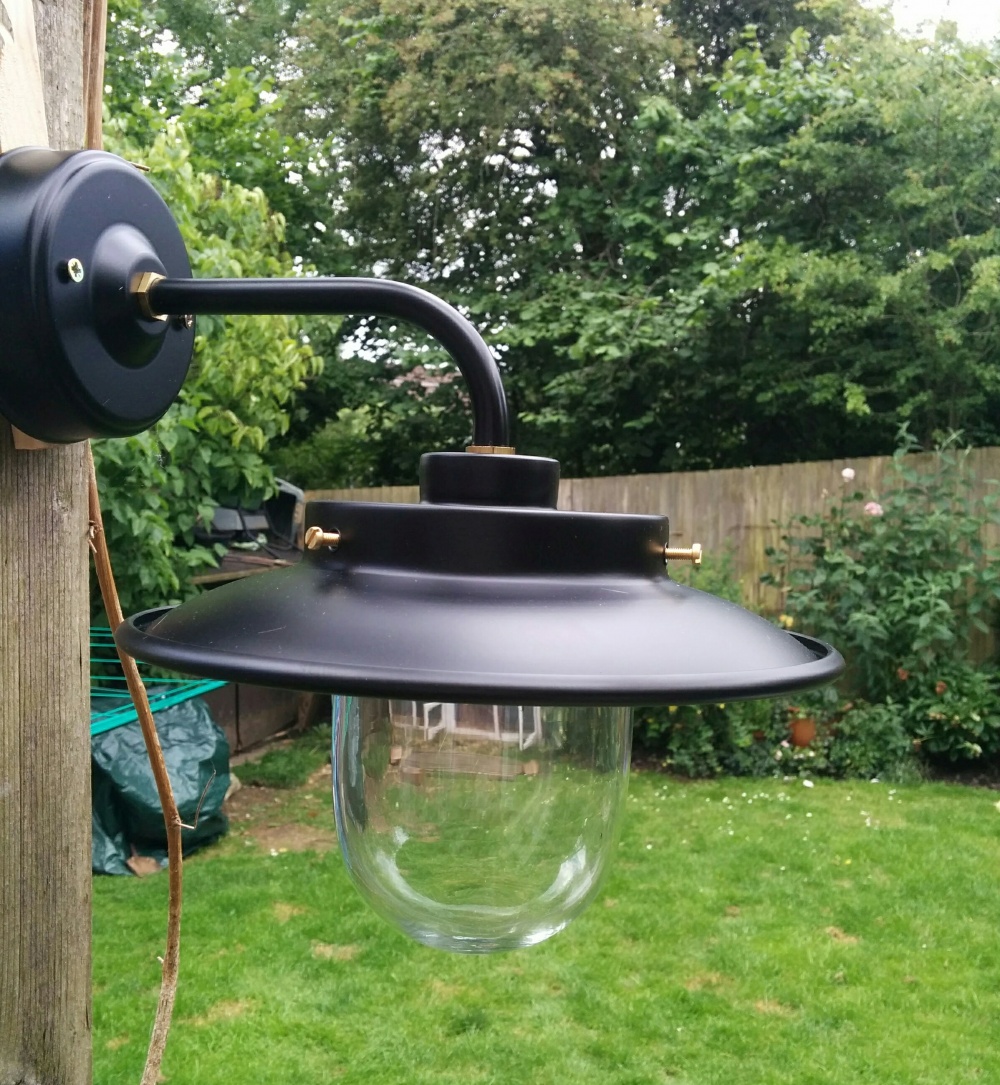 Image shows a sleek and stylish Burford Outdoor Wall Light in satin matt black against a backdrop of a charming outdoor setting